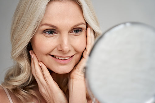 middle aged woman model touching face skin looking in mirror